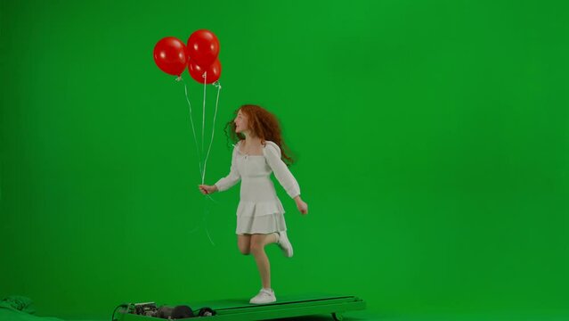 Redheaded little girl in white dress running with red balloons on green background of studio. Half turn. Concept of holiday, joy and fun.