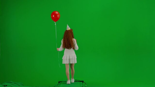 Redheaded little girl in white dress with a festive cap on her head and with a gift in her hands is walking with a red balloon. Green screen. Back view. Concept of holiday, joy and fun.