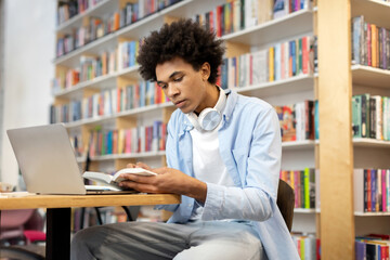 Focused black student guy sitting in library in front of laptop, diligently reading book, creating...
