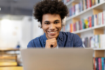 Black student guy, wearing headphones, looking at laptop screen with smile, sitting in library,...