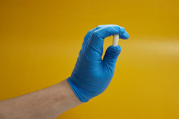 A hand in a blue medical glove holds a rectal suppository on a yellow background