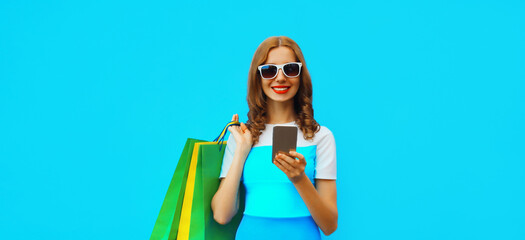 Beautiful happy young woman looking at mobile phone with shopping bag wearing summer dress on blue studio background - obrazy, fototapety, plakaty