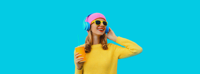 Portrait of modern cool young woman listening to music in headphones and drinking fresh juice wearing colorful pink hat, yellow sunglasses and sweater on blue studio background - obrazy, fototapety, plakaty