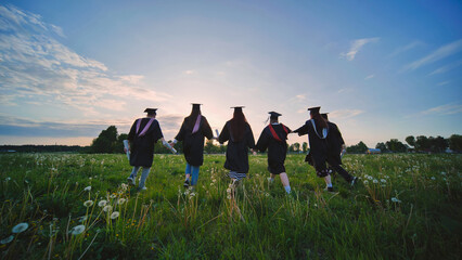 Six graduates in robes walk against the backdrop of the sunset.