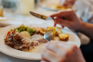 A person is eating dinner at the table using cutlery, close-up on hands and plate full of food