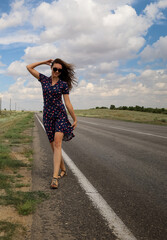 A young slender woman walking along the side of the highway against a cloudy sky 