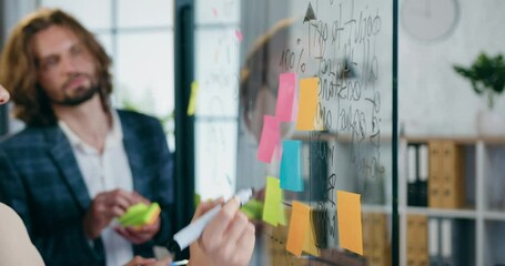Close up of professional man and woman colleagues analyzing business strategy of joint project using records on sticky notes attached to glass wall in office