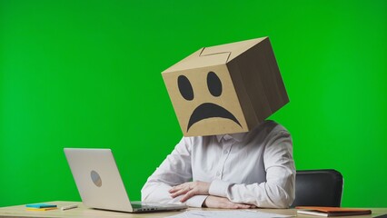 Woman in cardboard box with negative emoji on her head on green studio background. Employee sitting...