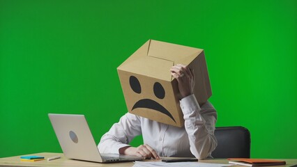 Woman in cardboard box with negative emoji on her head on green studio background. Employee sitting at desk, sighing frustrated and very tired. Business life and daily routine in the office.