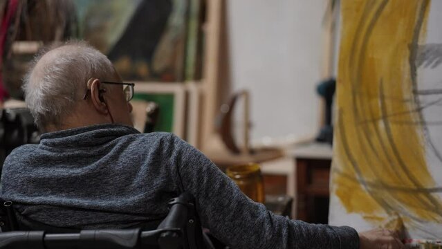 Senior artist on a wheelchair paints an oil painting on canvas in a studio