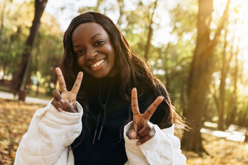 African american woman walking outdoor, portrait of young latin lady in warm sunny autumn park season, fall, yellow orange red leaves, dressed white jacket, having fun smiling laughing showing peace s