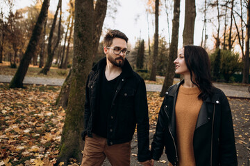 Heterosexual caucasian young loving couple walking outside in the city park in sunny weather, hugging smiling kissing laughing spending time together. Autumn, fall season, orange yellow red maple leav