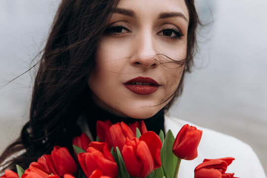 Attractive brunette woman walking on the beach shore in moody cloudy windy weather with bouquet of red tulips flowers, dressed in white suit jacket. International Women`s Day 8th March concept