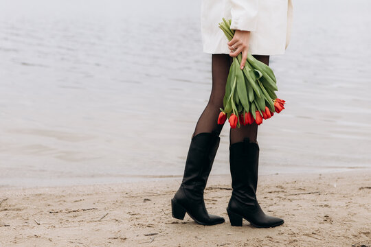 Attractive brunette woman walking on the beach shore in moody cloudy windy weather with bouquet of red tulips flowers, dressed in white suit jacket. International Women`s Day 8th March concept