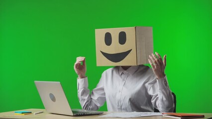 Woman in cardboard box with smiling emoji on her head on studio green background. The worker shows a thumbs up gesture and is happy about success, good news or winning.
