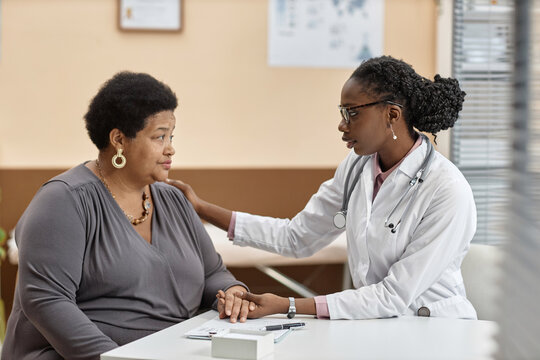 Side view of African American female physician calming mature woman patient while talking and holding her hand sitting at table in clinic office