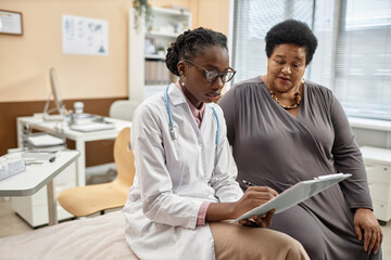 Medium full shot of Black woman physician in glasses holding clipboard and filling in medical record of senior female patient sitting in modern clinic office