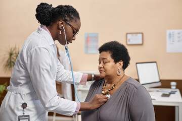 Young African American physician using stethoscope while asking senior female patient to make a...