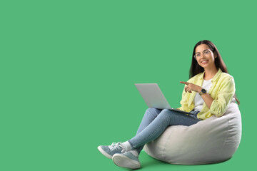 Young female programmer pointing at laptop while sitting on grey beanbag chair against green background