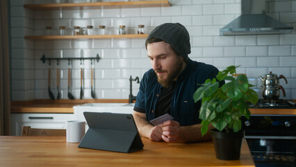 Young bearded man with beanie sitting in kitchen at home entering credit card number on tablet...
