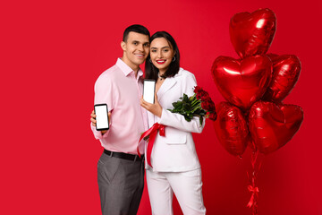 Young couple with mobile phones, bouquet of roses and heart-shaped air balloons on red background. Online dating