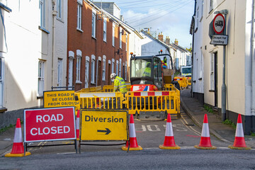 Road closed and road works image with road workers in fluorescent jackets operating machinery. Diversion sign in place. 