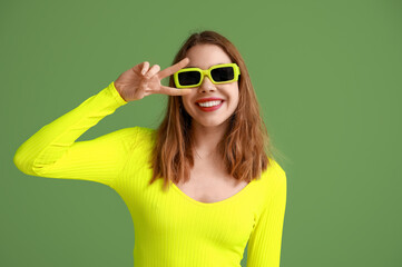 Pretty young woman in sunglasses showing peace gesture on green background
