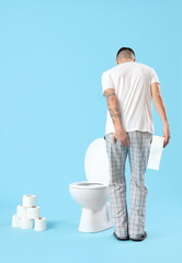 Young man with paper roll and toilet bowl on blue background, back view. Hemorrhoids concept