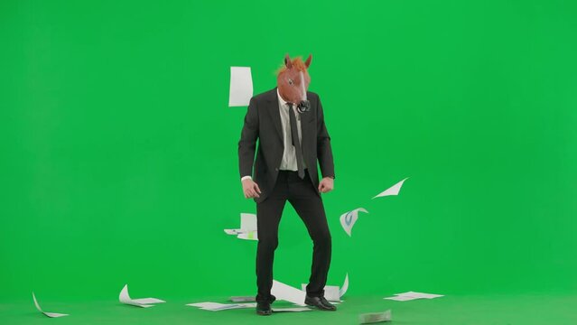 A man in a business suit with a horse head mask on a green studio background. The businessman looks through the documants, scatters them and begins to dance merrily. Hard office work concept.
