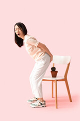 Young Asian woman with hemorrhoids and cactus on chair against pink background