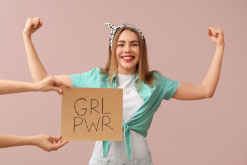 Female hands holding card with text GIRL POWER and happy young pin-up woman showing her muscles on pink background