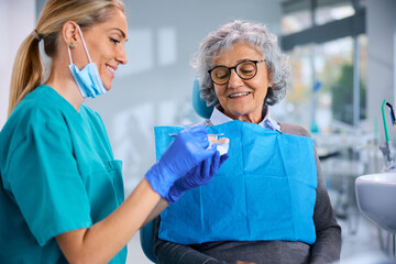 Female dentist explaining dental procedure to senior woman while using artificial denture at the...