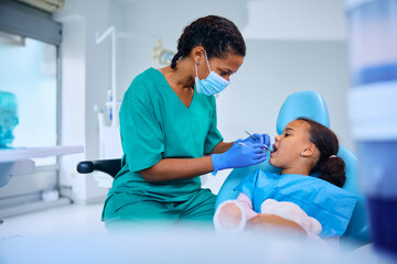 Black female dentist examining teeth of little girl at dental clinic.