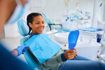 Happy black woman looking her teeth in mirror during appointment at dentist's office.