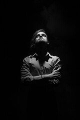 Fototapeta na wymiar Black and white portrait of a young man with a beard, standing on a black background in a ray of light, crossing his arms over his chest and looking up