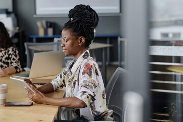 Side view shot of young black girl with dreadlocks working on tablet sitting at office desk