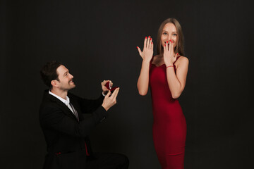 Beautiful romantic couple isolated on black background. Handsome man in suit is making proposal to his attractive young woman in red dress. Couple in love. Happy Saint Valentine's Day