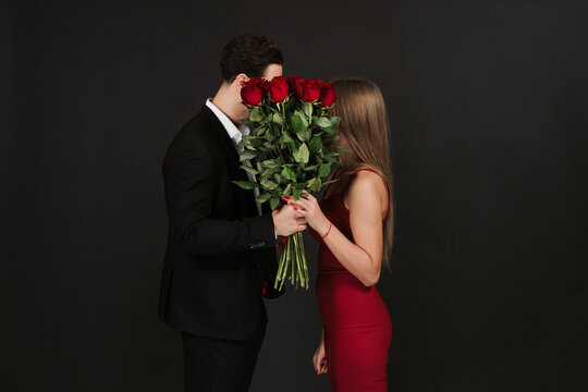 Beautiful romantic couple isolated on grey background. Attractive young woman in dress holding red roses and handsome man in suit are kissing. Happy Saint Valentine's Day