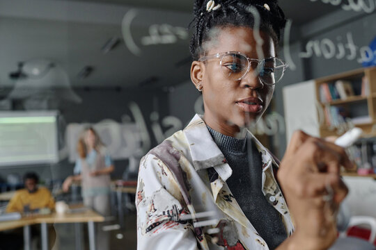 Female coder thoughtfully writing on glass wall in office, her colleagues working on background