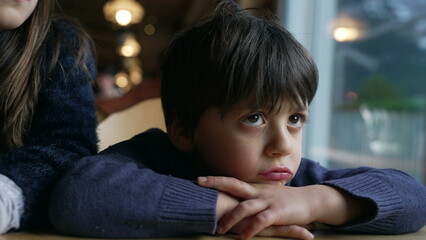 Bored small boy complaining, sulking child feeling annoyed leaning on restaurant table, closeup kid face