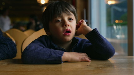 Bored small boy yawning while waiting at restaurant table by window. Child feeling boredom in authentic lifestyle moment awaiting for food to arrive