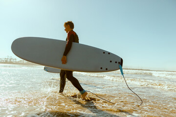 Back view of male surfer in wetsuit with his surfboard entering the sea. Surfing and ocean