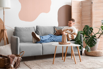 Young man reading book on sofa in living room - obrazy, fototapety, plakaty