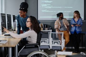 Black woman helping her colleague in wheelchair with code writing, coworkers standing on background