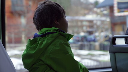 Obraz premium One small boy seated by bus window wearing green raincoat looking at view pass by inside transportation in motion