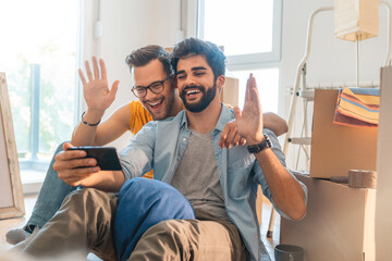 Joyous young gay couple making video call via mobile phone with unpacked boxes during relocation. Smiling happy homosexual couple waving hand during video call. - obrazy, fototapety, plakaty