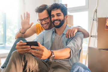 Young cheerful gay couple using mobile phone for video call waving hand sitting on floor with cardboard boxes during relocation. - obrazy, fototapety, plakaty