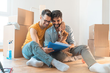 Happy gay couple smiling while cuddling dog and reading documents surrounded by cardboard boxes....