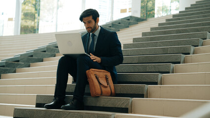 Skilled business man working by using laptop while sitting at stairs. Professional project manager...