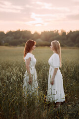 The image depicts two women wearing white dresses standing in a field of tall grass with a cloudy...